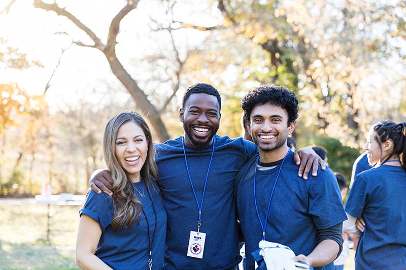 Trois personnes portent le même t-shirt bleu et des cordons autour du cou. Ils sourient pour une photo de groupe.  Il s'agit d'un cadre extérieur avec des arbres en arrière-plan.