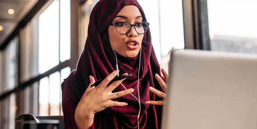 Une femme portant des lunettes et un hijab violet semble animée lors d'un appel vidéo sur son ordinateur portable.
