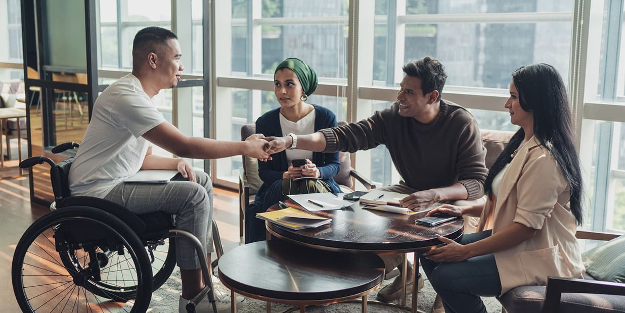 Un groupe de personnes s'assoit à une table et interagit. Des blocs-notes et des stylos sont posés sur la table. Deux hommes se serrent la main de l'autre côté de la table, l'un d'eux étant assis dans un fauteuil roulant.