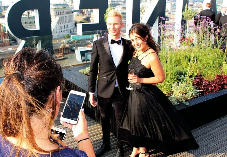 Deux personnes, habillées en tenue de gala, se trouvent sur un toit-terrasse. Une troisième personne les prend en photo avec son téléphone. Le temps est ensoleillé.