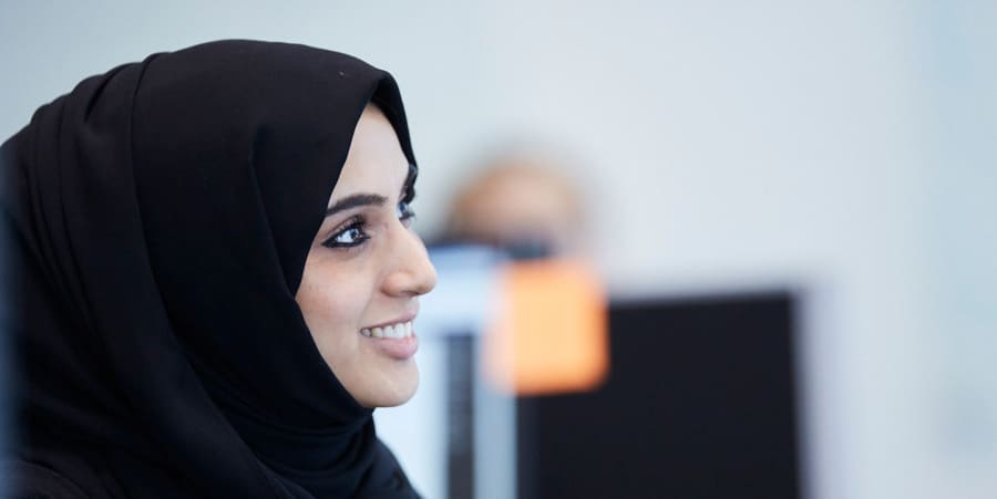 Une femme portant un hijab noir sourit et détourne son regard de la caméra. La photo est un angle latéral de son visage.