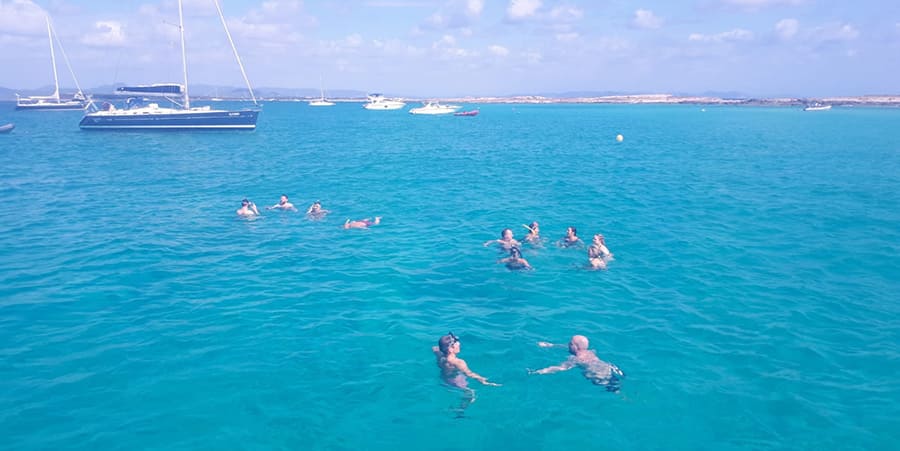 Un groupe de personnes profite d'une baignade dans la mer. On aperçoit des bateaux au loin.