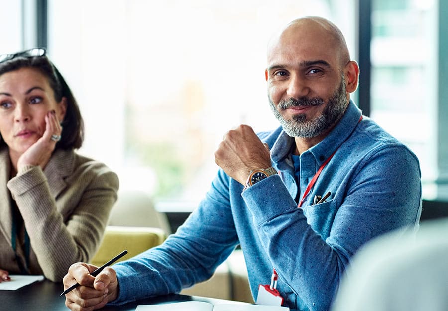 Un environnement professionnel avec un homme et une femme à une table. L'homme au centre de l'image porte une chemise bleue. Il y a un cahier devant lui et il tient un stylo. Il sourit.