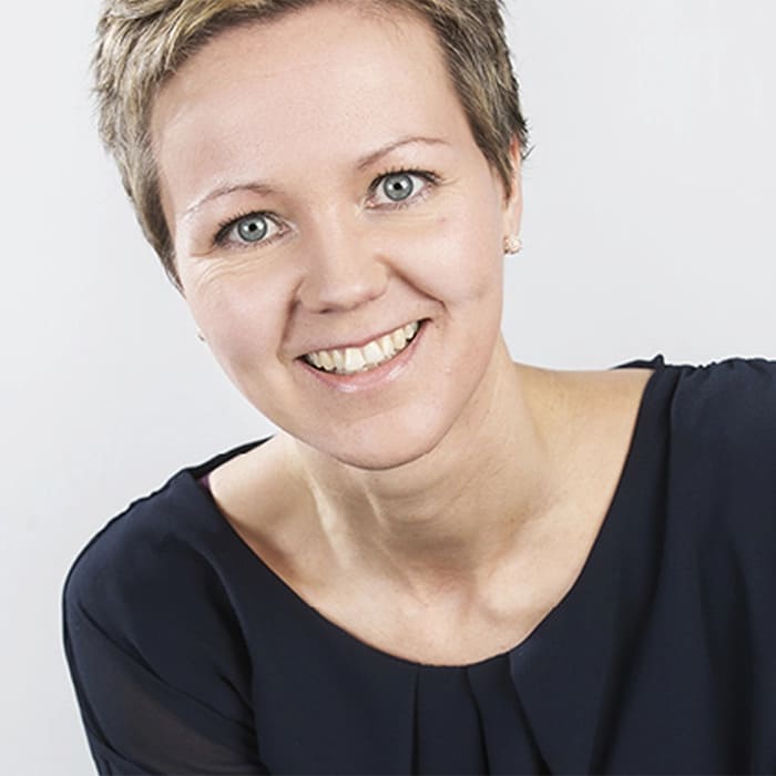 Une femme aux cheveux bruns courts sourit à la caméra. Il s'agit d'un studio.