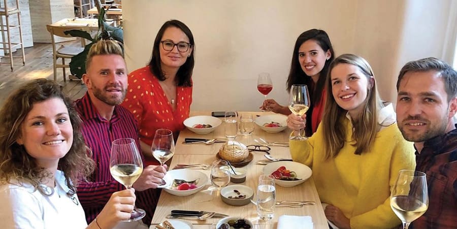 Six personnes sont assises à une table dans un restaurant. Ils tiennent un verre de vin et sourient à la caméra.