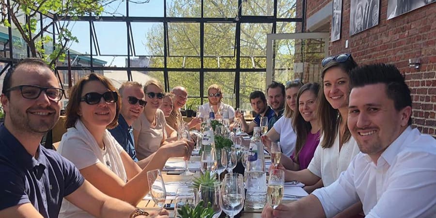 Un groupe d'environ 15 personnes est assis à une longue table et boit un verre. Ils sourient tous à la caméra. Ils portent des vêtements décontractés.