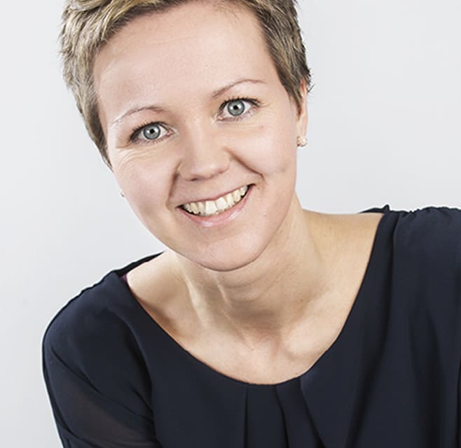 Une femme aux cheveux bruns courts sourit à la caméra. Il s'agit d'un studio.